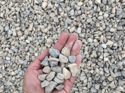 A hand holding three-quarter inch construction gravel above a bed of the same clean grey rock