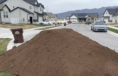 A long pile of screened topsoil dropped along a residential street, houses and mountains behind